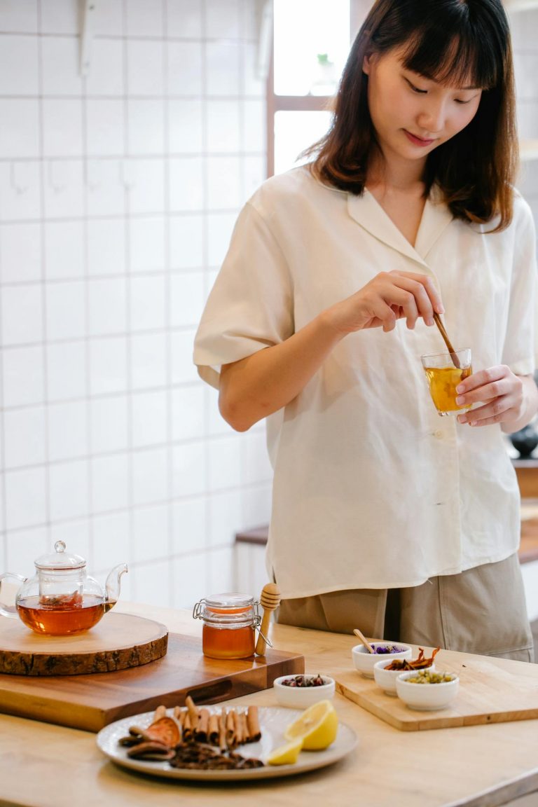 Asian woman stirring herbal tea with honey in a modern kitchen, surrounded by tea ingredients.