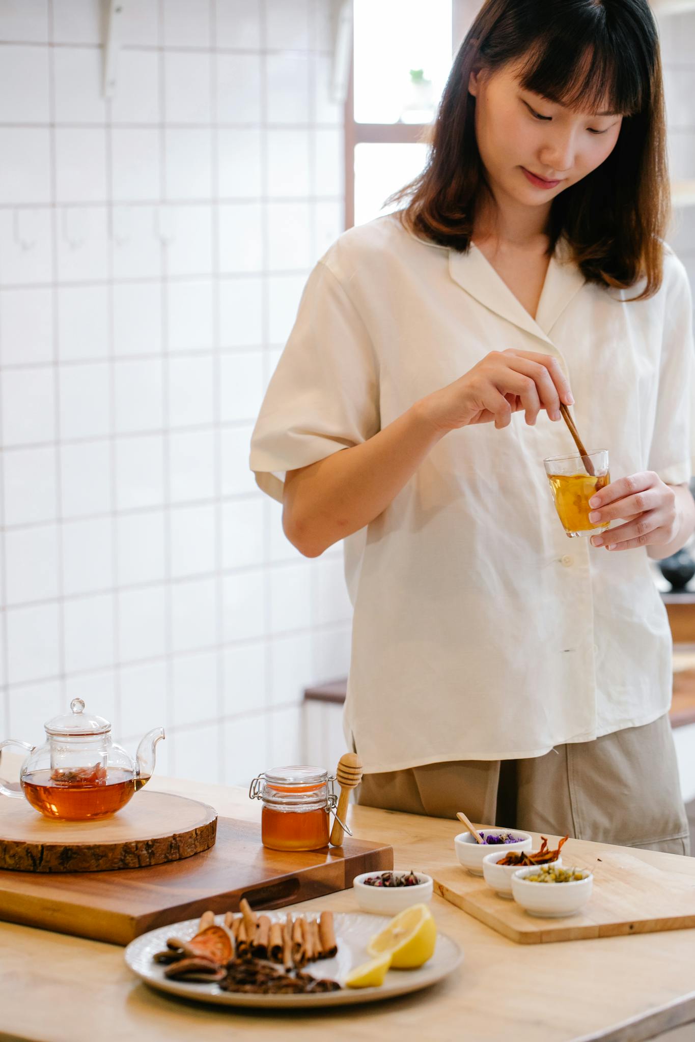Asian woman stirring herbal tea with honey in a modern kitchen, surrounded by tea ingredients.