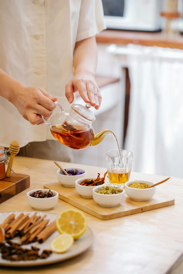 Person pouring herbal tea into a glass surrounded by spices and herbs on a kitchen table.